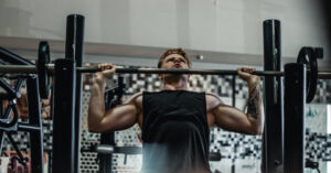man using a wide grip to overhead press a loaded barbell inside a squat rack at the gym